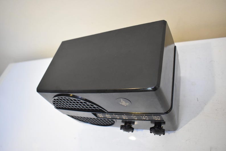 Puede incluir: Una radio negra vintage con forma rectangular redondeada. La radio tiene una rejilla de altavoz, un dial de sintonizaci&oacute;n y dos perillas de control. La cara de la radio muestra los n&uacute;meros de frecuencia. Un equipo de audio cl&aacute;sico.
