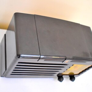 May include: A vintage, dark grey radio with a rectangular shape. The radio has a series of horizontal lines on the front, with two black knobs and a small, illuminated section. The radio is mounted on a white surface.