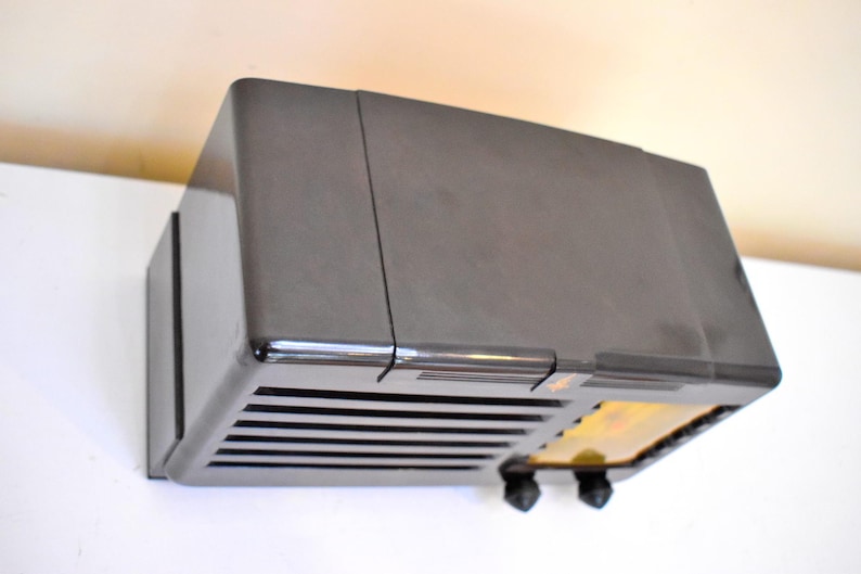 May include: A vintage, dark grey radio with a rectangular shape. The radio has a series of horizontal lines on the front, with two black knobs and a small, illuminated section. The radio is mounted on a white surface.
