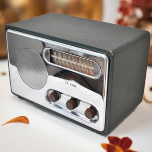 May include: Vintage black and silver radio with a speaker, tuning dial, and three brown knobs. The radio has a rectangular shape and a chrome-colored face. The radio is a retro style.