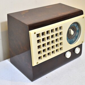 May include: A vintage brown wooden radio with a white plastic face and a blue dial. The radio has a grid-like speaker and two white knobs.