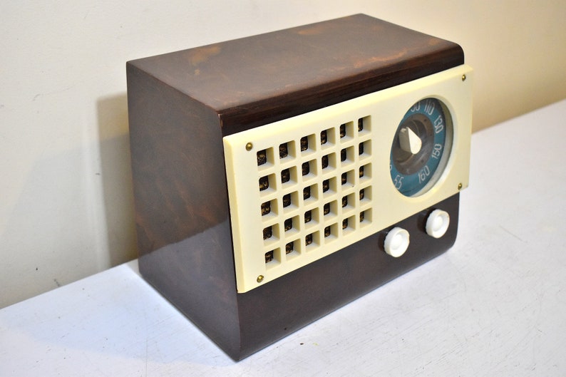 May include: A vintage brown wooden radio with a white plastic face and a blue dial. The radio has a grid-like speaker and two white knobs.