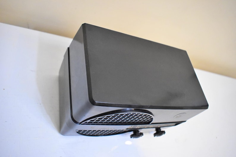 Puede incluir: Un dispositivo electr&oacute;nico rectangular gris oscuro con una parte superior ligeramente inclinada. El dispositivo tiene una rejilla de malla en la parte delantera y dos perillas negras. El objeto est&aacute; sobre una superficie blanca y una pared beige.