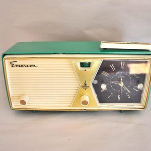 May include: A vintage Emerson clock radio. It has a cream-coloured front, a green top, and gold accents. The clock face is black with white numerals and hands. Features two cream-coloured knobs and the Emerson logo.