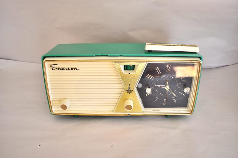 May include: A vintage Emerson clock radio. It has a cream-coloured front, a green top, and gold accents. The clock face is black with white numerals and hands. Features two cream-coloured knobs and the Emerson logo.