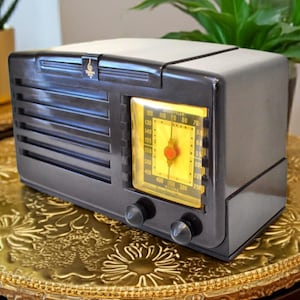 May include: A retro black radio featuring a yellow illuminated dial and two silver control knobs. The radio, with its horizontal grill lines, sits on a gold, ornate table. A small gold emblem is on top.