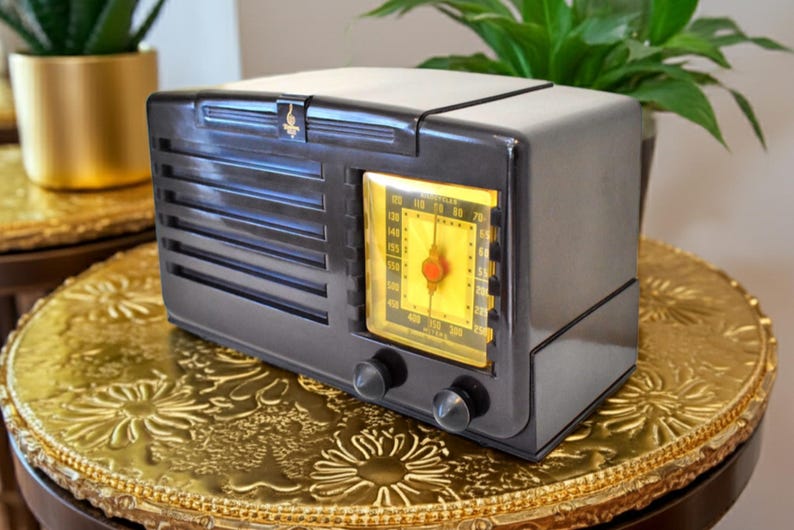 May include: A retro black radio featuring a yellow illuminated dial and two silver control knobs. The radio, with its horizontal grill lines, sits on a gold, ornate table. A small gold emblem is on top.
