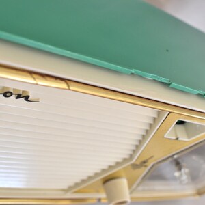 May include: A close-up of a vintage Emerson appliance. The appliance has a mint green top, a cream-coloured body, and gold trim. The name "Emerson" is printed in black script. The design is reminiscent of mid-century modern aesthetics.