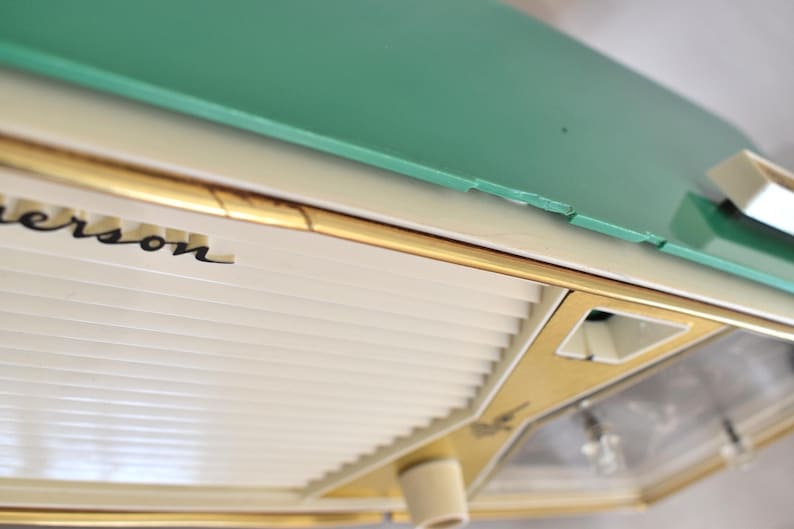 May include: A close-up of a vintage Emerson appliance. The appliance has a mint green top, a cream-coloured body, and gold trim. The name "Emerson" is printed in black script. The design is reminiscent of mid-century modern aesthetics.