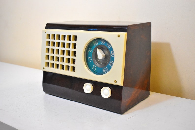 May include: A vintage brown and cream colored radio with a blue dial and a white knob. The radio has a speaker grille on the front and two knobs on the bottom.
