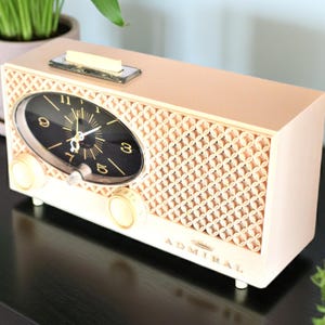 May include: A vintage-style clock radio in cream and peach. The clock face is black with gold numerals and hands. The speaker grill has a geometric pattern. The word "Admiral" is printed on the front.