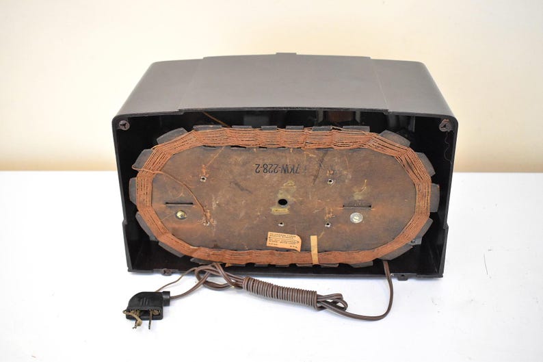 May include: A vintage black radio with a brown speaker and a power cord. The radio's back panel is visible, showing the speaker's components and a label with the text "7KW 228 2". The radio is on a white surface.