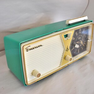 May include: A retro Emerson clock radio in teal and cream. The clock face is black with gold detailing. The radio features a cream speaker grill and gold trim. The Emerson logo is displayed on the front.