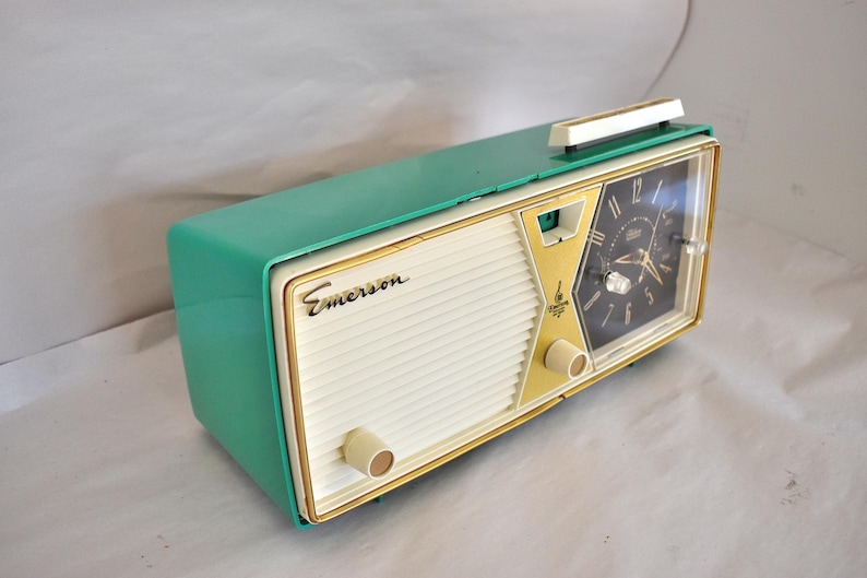 May include: A retro Emerson clock radio in teal and cream. The clock face is black with gold detailing. The radio features a cream speaker grill and gold trim. The Emerson logo is displayed on the front.