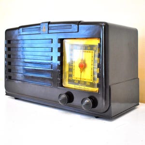 May include: A vintage black radio with a yellow dial and a red needle. The radio has a rectangular shape with horizontal lines on the front and two knobs. The radio is on a white surface.