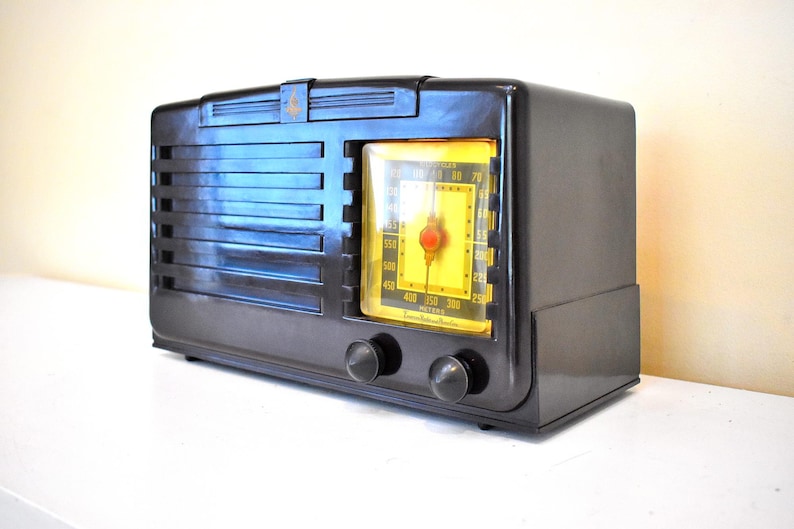 May include: A vintage black radio with a yellow dial and a red needle. The radio has a rectangular shape with horizontal lines on the front and two knobs. The radio is on a white surface.