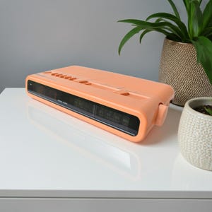 May include: A peach-colored vintage digital clock radio with a black display screen. The clock has several orange buttons on top and a tuning dial on the side. The clock is on a white surface.