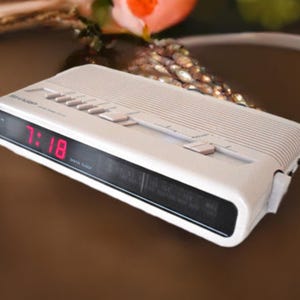 May include: White digital clock radio with a red digital display showing the time 7:18. The clock radio has several buttons and a black speaker grille.