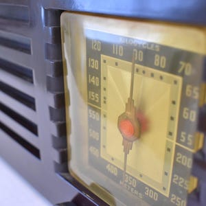 May include: A close-up of a vintage brown radio with a yellowed dial. The dial shows numbers and a needle, with the text "KILOCYCLES" and "METERS". The radio has a dark brown, rectangular body with vertical slats.
