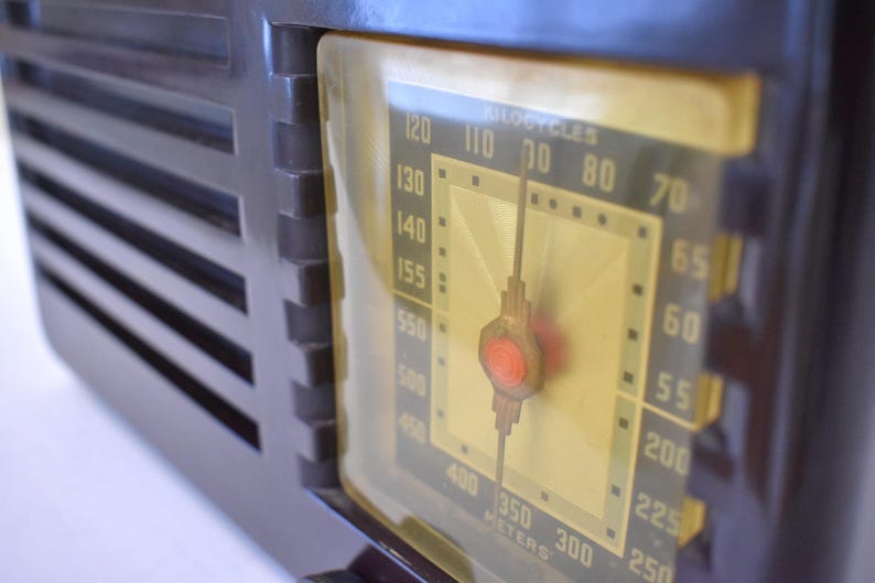 May include: A close-up of a vintage brown radio with a yellowed dial. The dial shows numbers and a needle, with the text "KILOCYCLES" and "METERS". The radio has a dark brown, rectangular body with vertical slats.