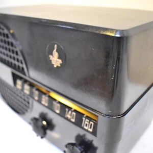 Puede incluir: Una radio negra vintage con un emblema dorado y un dial de sintonizaci&oacute;n. La radio tiene forma rectangular con bordes redondeados y una rejilla de altavoz en el lado izquierdo. El dial muestra n&uacute;meros del 60 al 160.