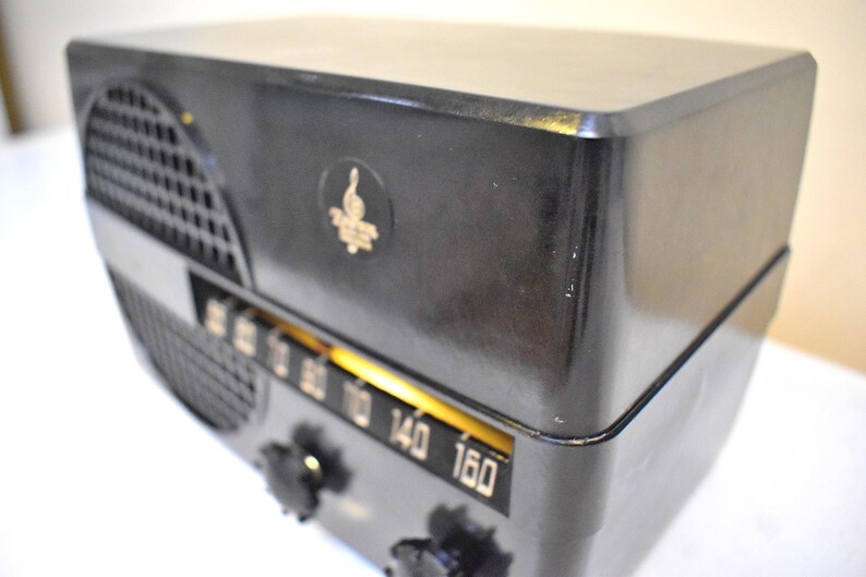 Puede incluir: Una radio negra vintage con un emblema dorado y un dial de sintonizaci&oacute;n. La radio tiene forma rectangular con bordes redondeados y una rejilla de altavoz en el lado izquierdo. El dial muestra n&uacute;meros del 60 al 160.