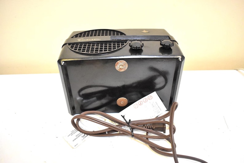 Puede incluir: Una radio Emerson negra vintage con una rejilla de altavoz, dos perillas y un cable de alimentaci&oacute;n. La radio tiene forma rectangular y un peque&ntilde;o emblema en la parte superior. Una etiqueta de papel est&aacute; adherida al cable de alimentaci&oacute;n.