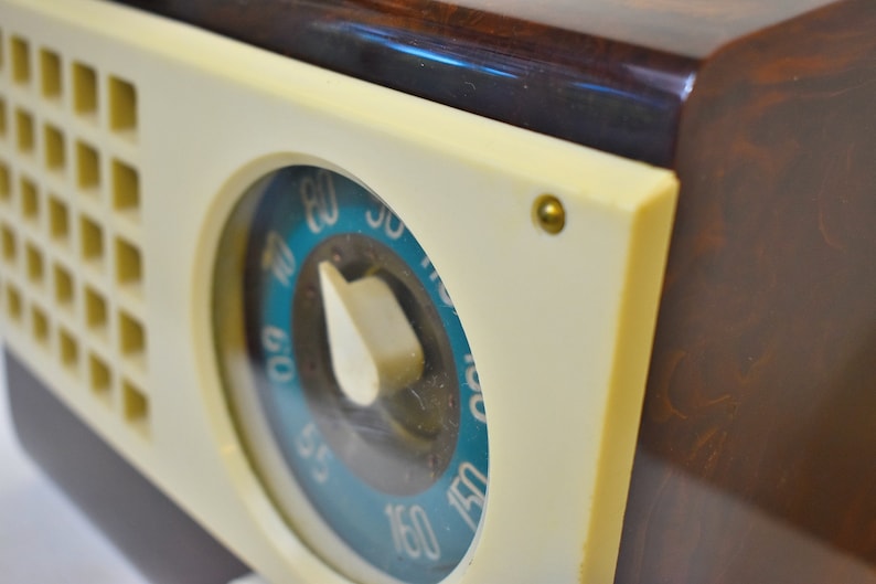 May include: Close-up of a vintage radio with a white face and a blue and white dial. The dial has numbers from 55 to 150. The radio has a brown wooden case.