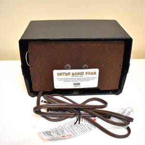 Puede incluir: Una radio retro negra con un panel de altavoz marr&oacute;n y un cable de alimentaci&oacute;n. La radio tiene forma rectangular con una etiqueta que dice "Retro Radio Farm". El cable de alimentaci&oacute;n es marr&oacute;n y tiene una etiqueta.