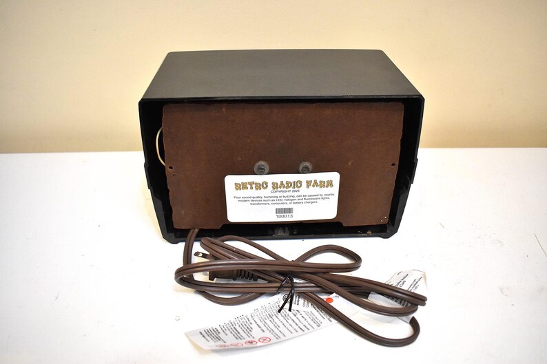 Puede incluir: Una radio retro negra con un panel de altavoz marr&oacute;n y un cable de alimentaci&oacute;n. La radio tiene forma rectangular con una etiqueta que dice "Retro Radio Farm". El cable de alimentaci&oacute;n es marr&oacute;n y tiene una etiqueta.