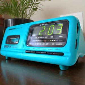 May include: A vintage turquoise digital alarm clock with a cassette player. The time displayed is 2:03 in green digits. It has an AM/FM radio dial and black control buttons. The clock is on a dark wooden surface.