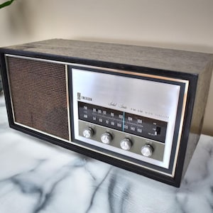 May include: A vintage Emerson Solid State AM/FM radio with a black wood cabinet and a silver faceplate. The radio has a speaker grille, tuning knobs, and a dial with numbers for AM and FM frequencies.