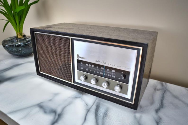 May include: A vintage Emerson Solid State AM/FM radio with a black wood cabinet and a silver faceplate. The radio has a speaker grille, tuning knobs, and a dial with numbers for AM and FM frequencies.