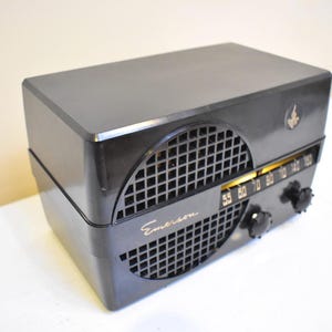 Puede incluir: Una radio Emerson negra vintage con una cubierta de rejilla para el altavoz y letras doradas. La radio tiene un dial de sintonizaci&oacute;n con n&uacute;meros y dos perillas negras. Un peque&ntilde;o emblema est&aacute; en la esquina superior derecha.