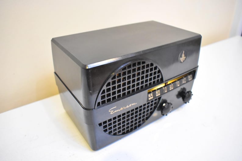 Puede incluir: Una radio Emerson negra vintage con una cubierta de rejilla para el altavoz y letras doradas. La radio tiene un dial de sintonizaci&oacute;n con n&uacute;meros y dos perillas negras. Un peque&ntilde;o emblema est&aacute; en la esquina superior derecha.