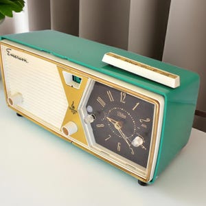 May include: A vintage Emerson clock radio in a retro design. The clock radio has a teal green top and sides, with a cream-coloured front panel and gold accents. The clock face is black with gold numerals and hands. A white bar sits on top.