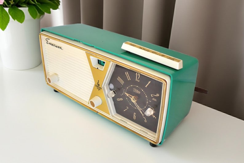 May include: A vintage Emerson clock radio in a retro design. The clock radio has a teal green top and sides, with a cream-coloured front panel and gold accents. The clock face is black with gold numerals and hands. A white bar sits on top.
