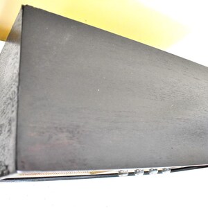 May include: A black wooden box with a textured surface. The box has a small, rectangular opening on the front with a row of silver knobs.