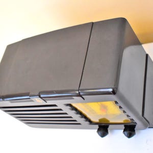May include: A vintage, dark brown radio with a rectangular shape. The radio has a speaker grille, two black knobs, and a yellow display. The radio is mounted on a white wall.