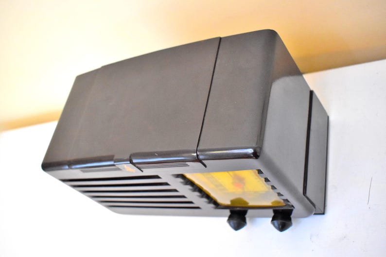 May include: A vintage, dark brown radio with a rectangular shape. The radio has a speaker grille, two black knobs, and a yellow display. The radio is mounted on a white wall.