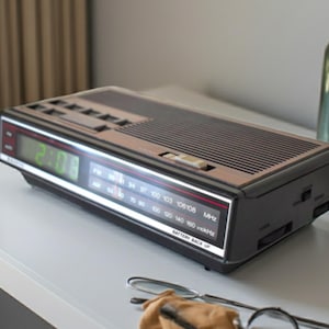 Könnte beinhalten: Ein schwarzer und brauner digitaler Wecker mit einer grünen LED-Anzeige, die die Uhrzeit anzeigt. Die Uhr hat ein AM/FM-Radio und eine Batterie-Backup-Funktion. Die Uhr steht auf einer weißen Oberfläche.
