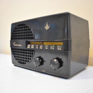 Puede incluir: Radio Emerson negra vintage con rejilla de altavoz, dial de sintonizaci&oacute;n y dos perillas. El dial de la radio muestra n&uacute;meros del 55 al 160. Hay un emblema dorado en la parte superior. La radio est&aacute; sobre una superficie blanca.