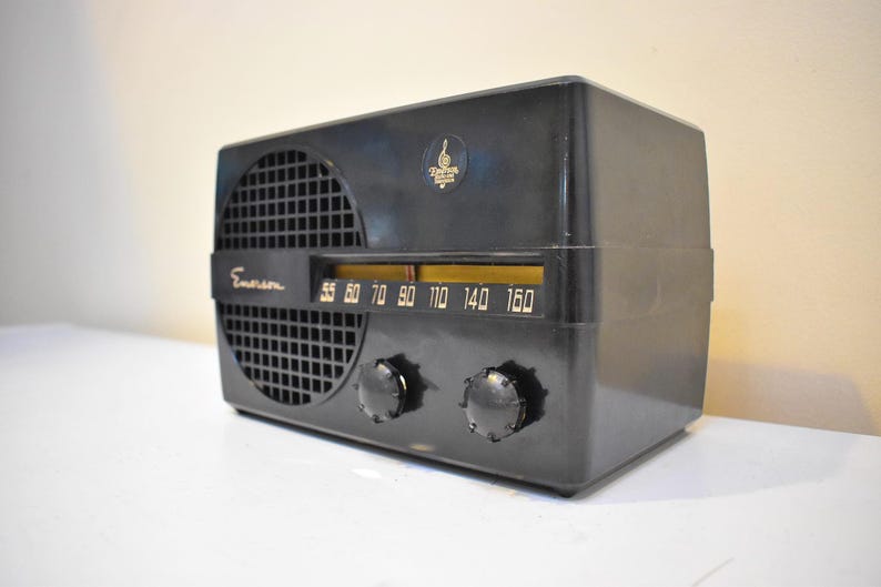 Puede incluir: Radio Emerson negra vintage con rejilla de altavoz, dial de sintonizaci&oacute;n y dos perillas. El dial de la radio muestra n&uacute;meros del 55 al 160. Hay un emblema dorado en la parte superior. La radio est&aacute; sobre una superficie blanca.