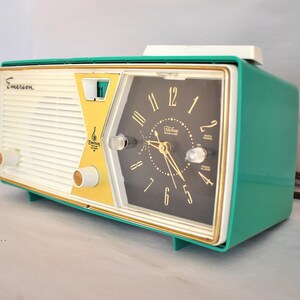 May include: A retro Emerson clock radio in teal, cream, and gold. The clock face is black with gold hands and numerals. The radio has a vintage design with the Emerson logo on the front.