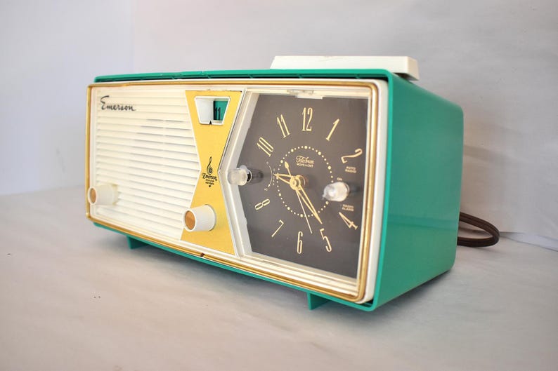 May include: A retro Emerson clock radio in teal, cream, and gold. The clock face is black with gold hands and numerals. The radio has a vintage design with the Emerson logo on the front.