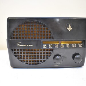 Puede incluir: Una radio Emerson negra vintage con una rejilla de altavoz dorada. La radio tiene un dial de sintonizaci&oacute;n con n&uacute;meros y dos perillas. El logotipo de Emerson es visible en la parte delantera.