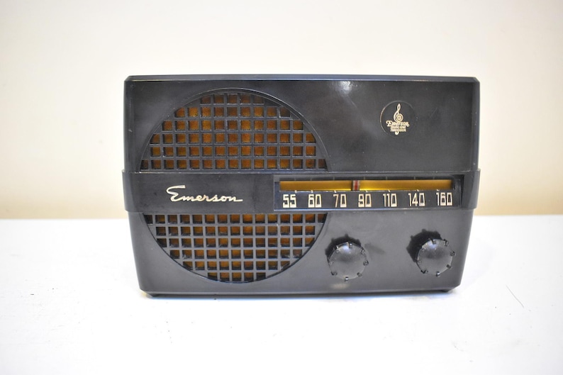 Puede incluir: Una radio Emerson negra vintage con una rejilla de altavoz dorada. La radio tiene un dial de sintonizaci&oacute;n con n&uacute;meros y dos perillas. El logotipo de Emerson es visible en la parte delantera.