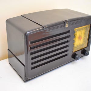 May include: A vintage, dark grey radio with a glossy finish. The radio has a rectangular design with horizontal vent lines and a yellow dial. Two black knobs are visible on the front. A small gold emblem is on the top.