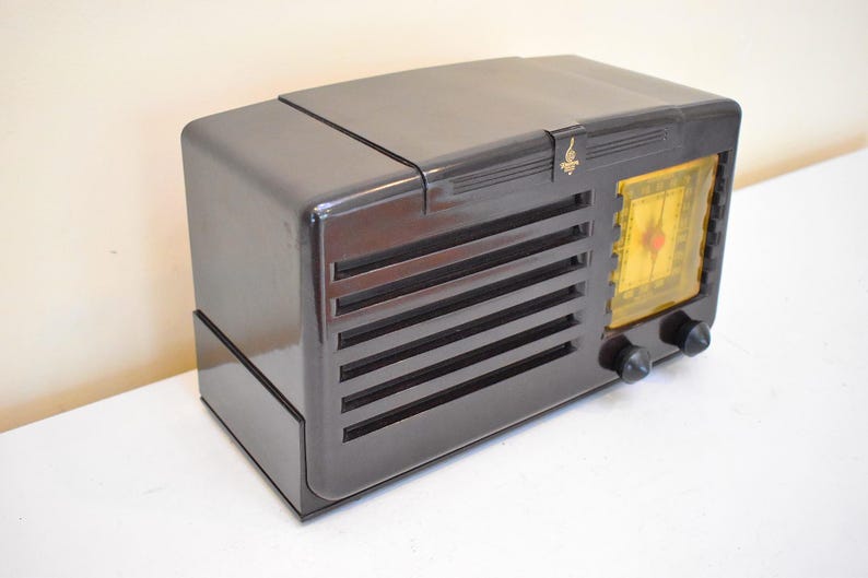 May include: A vintage, dark grey radio with a glossy finish. The radio has a rectangular design with horizontal vent lines and a yellow dial. Two black knobs are visible on the front. A small gold emblem is on the top.