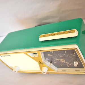 May include: A retro green and cream alarm clock with a gold-coloured trim. The clock face is black with gold hands and numerals. The top of the clock has a white panel with the words "Sleep Saver" and "Dose Alarm".